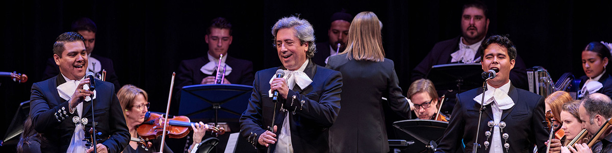 Three mariachi vocalists performing with a full orchestra onstage, led by a conductor, during the Balboa 100 Gala.