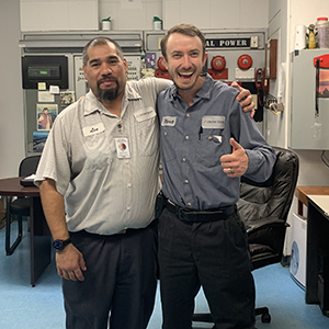 Two engineering employees standing together in a workshop office, smiling as one gives a thumbs-up.