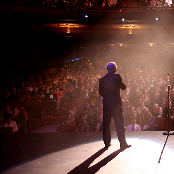 Behind-the-scenes stage view of a speaker at a microphone addressing a packed Balboa Theater audience.