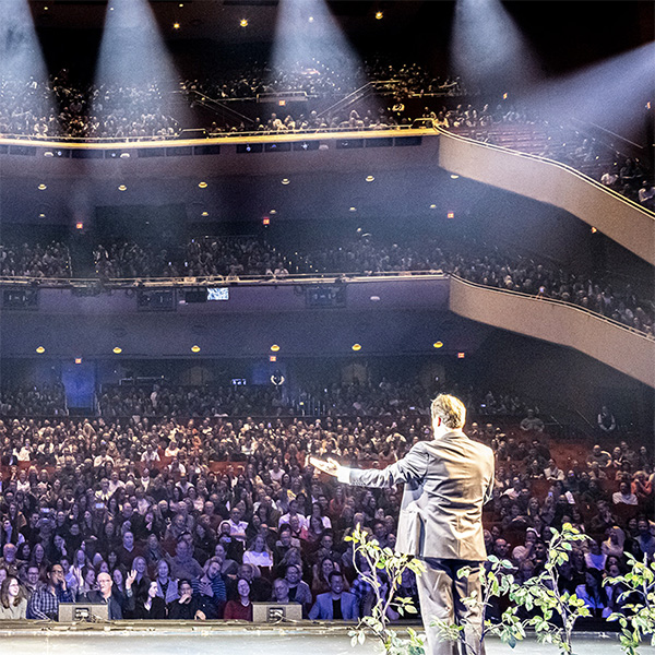 Behind-the-scenes stage view of a speaker at a microphone addressing a packed Civic Theater audience.