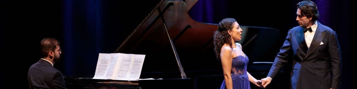 On stage, a man and woman singing to each other with a man playing the piano to their left.